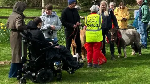A group of people in hiking gear some on bikes and one wheelchair user, a dog and two small ponies on a green field beside a wooden fence with a stile and a trail sign 