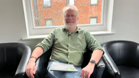 A worker from drug and alcohol service Via, which supports young people experiencing ketamine harms. He is sitting in a leather black armchair with a window behind him through which you can see a redbrick building. He has a white beard and hair and is wearing glasses, a green buttoned-up shirt and blue jeans.