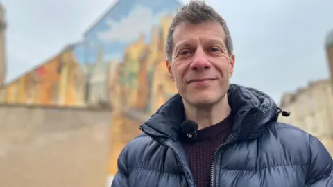Emma Baugh/BBC A man, with short grey hair, wearing a dark blue puffer jacket, standing in front of a street mural (which is blurred), smiling for the camera.