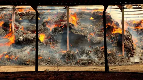 South Yorkshire Fire and Rescue Service The interior of a large open-sided structure containing substantial piles of hay that are actively burning. The scene is framed by a series of dark vertical support columns and a roof overhead. The hay appears heavily charred, with deep black and grey surfaces broken up by numerous pockets of bright orange and yellow flames. Fire is visible in many different locations across the piles.