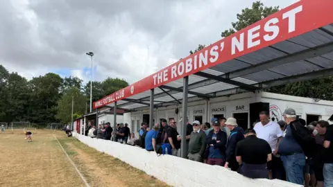 A photo of football stands at the top it says THE ROBINS' NEST in red with white writing. There are around 50 people standing and watching the football. 