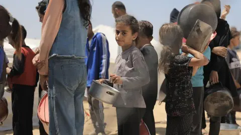 Getty Images Palestinians, including children wait to receive hot meals ,distributed by a Palestinian charity to displaced Palestinians living in tents in western Gaza City