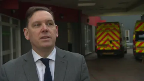 Man in suit with navy tie and white shirt stands in front of ambulance outside A&E