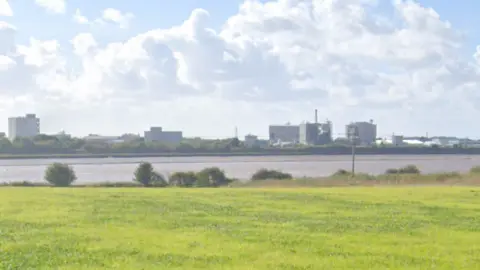 Google The Hillhouse chemical plant, a large, industrial complex consisting of several large grey buildings, sit on the opposite bank of the River Wyre. In the foreground is a field.