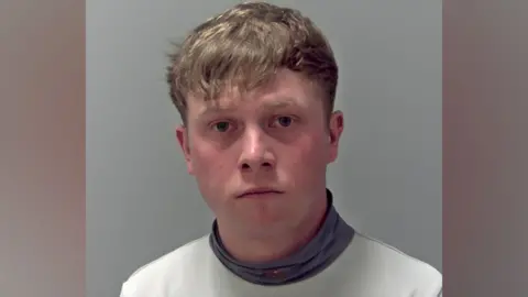 A police custody photo of Levi Williams. He has light brown hair which over hangs on to his forehead. He wears a white top with a grey turtleneck. 