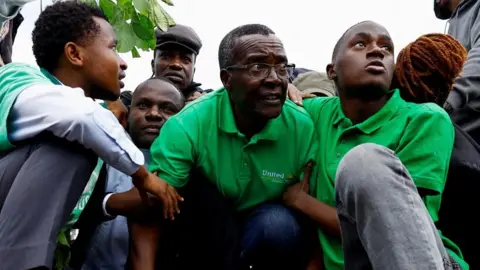 David Maraga in a green T-shirt crouches on the floor with other protesters. He is flanked by two others who are holding his arms.