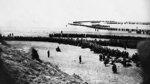 A black and white image of a long line of soldiers on a beach. 