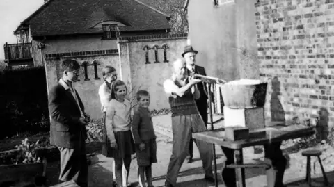 Wallis Family A black and white image of a family stood in a garden firing a catapult at a water bucket.