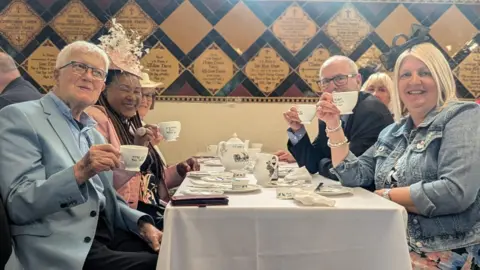 Stoke-on-Trent City Council A group of people sat either side of a table with a white cloth and tea set on top. They are holding up their mugs and smiling.