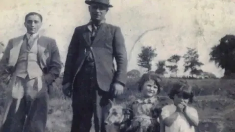 An old black and white photograph showing two men dressed in suits, with one wearing a hat, standing next to a dog and two young children. Trees and fields can be seen in the background.