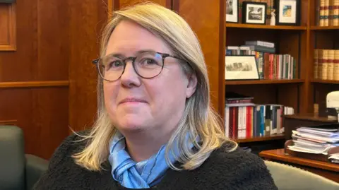 The Lady Chief Justice has shoulder length blonde hair, she is wearing brown round glasses, a blue scarf and a black jumper. Behind her is a mahogany wall and shelves with books and framed pictures on them. 