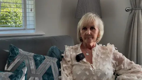 A woman at on a sofa wearing a white floral blouse looking to the side of the camera with a serious facial expression