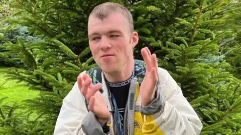 A person wearing a light-coloured jacket stands outdoors in front of dense green fir trees, holding their hands raised as if they are clapping.
