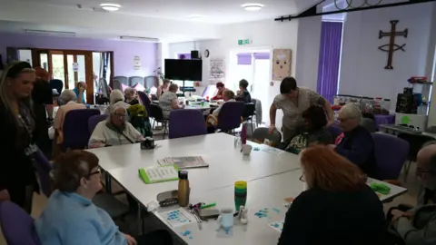 BBC/Nick Gemzoe A group of people sitting at tables chatting and playing bingo at one of the Trinity Network sessions.