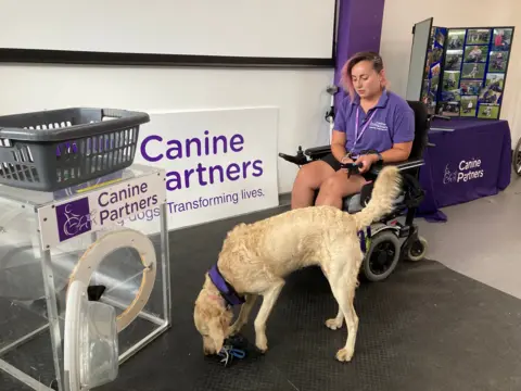A dog learning to load a prop washing machine made from perspex under instruction from a woman in a wheelchair