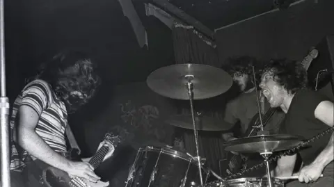 Getty Images Rory Gallagher in striped t-shirt, with John Wilson on drums, and Richard McCracken, performing at a gig in Germany