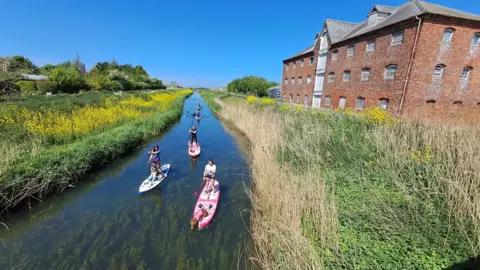 LNT Four paddleboarders and a kayaker paddling in a canal, which is flanked by marshland. On the right is a large red-brick three-storey building. The paddlers are moving towards the camera.