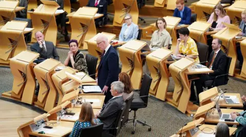 John Swinney stands next to a wooden desk in Holyrood surrounded by MSPs