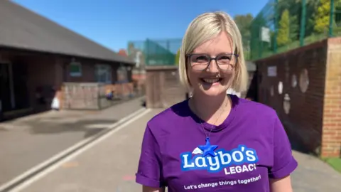 BBC / Simon Spark Emily Kitchener. She has short blond hair and is wearing glasses and a purple T-shirt with the words Laybo's Legacy - let's change things together.