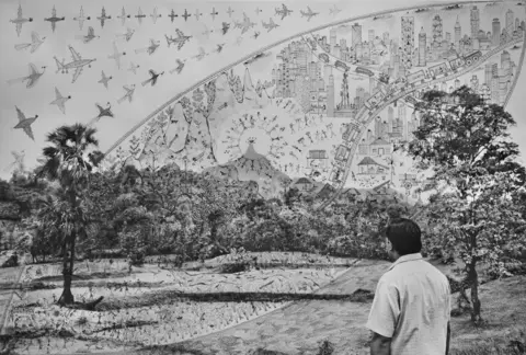 Gauri Gill and Rajesh Vangad Black and white photograph of a person with their back turned, looking out to a forest, layered with Warli drawings over the image