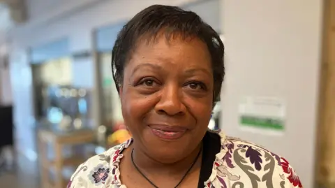 A woman with short black hair, she is looking into the camera and smiling, and wearing a white floral top. 