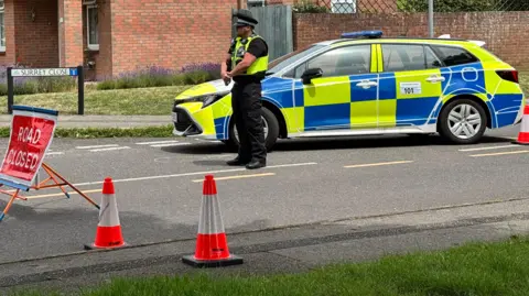 Police officer standing behind cordon on a road with police car behind and homes in the background.