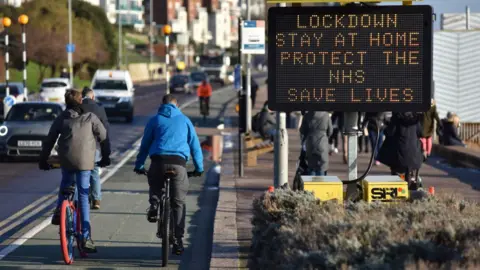 Getty Images Cyclists ride past a digital public safety notice saying "Lockdown, Stay At Home, Protect The NHS, Save Lives" along the seafront at Westcliff, in Southend on 6 January