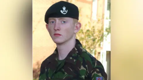 Cyrus Thatcher is wearing his army uniform. He has short, ginger hair and is stood expressionless.