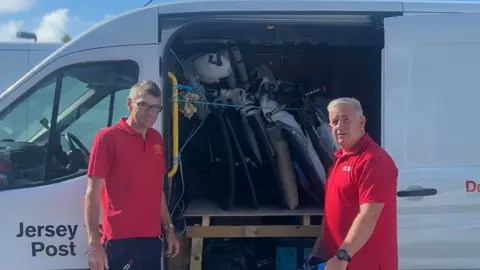 Alan and Toby stood in front of the Jersey Post van door. It is filled with equipment and Alan is putting a bag into it.