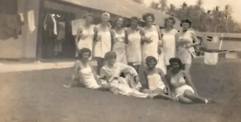 Gillian Edwards An old black and white image of about a dozen young women in white negligees and underwear posed on a lawn in front of a low makeshift building - some are sitting and some standing behind, all smiling to camera. Clothes are drying on a washing line in the background