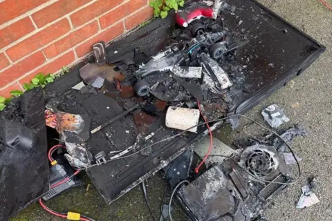 Sherianne Wood A tray which has been burnt, with various wires and electrics and a car, led on the ground next to a brick wall.