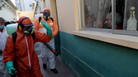 Reuters A worker wearing protective gear waves to a woman while using disinfectant to clean outside of a house in Santiago, Chile