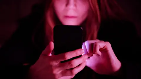 Getty Images A woman is bathed in pink light as she looks at her phone in her hand