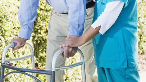 Getty Images Carer supporting patient walking with a frame