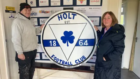 BBC Nigel Tripp and Joy Bloomfield standing either side of football club sign