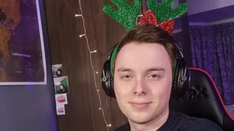 EnDjinn A man sits facing the camera while smiling wearing headphones and festive sparkly green reindeer antlers