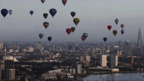 Hot air balloons soar above central London with the River Thames in the foreground and the Shard, Eye and other buildings are in the background