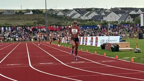 Manx Radio Sport Christa Cain wears a red crop top, black shorts and track spikes. She crosses the finish line on an orange 400m track. There are other runners in the distance, and large crowds of people watch on.