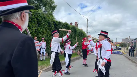 A Morris dancing troupe waves their batons in the air, dressed in traditional clothing. Bystanders watch the dancing on the village street, which is decorated in Union Jack bunting.