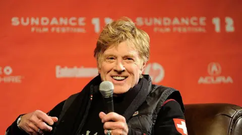 Robert Redford, a handsome older man with blonde hair smiles broadly while speaking on stage at a Sundance Festival Event. The festival's logo is on a backdrop behind him he sits in a brown leather chair, while wearing a black jumper with a Swiss flag on the arm, and a warm puffa gilet in black.