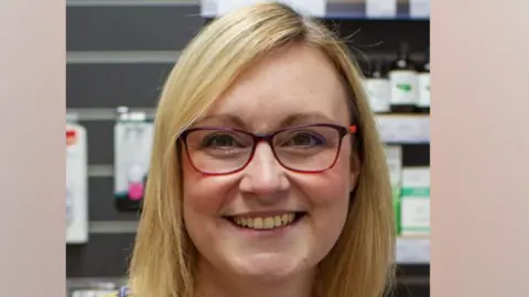 Monkbar Pharmacy A woman with blonde hair and wearing glasses smiles at the camera, in a pharmacy.