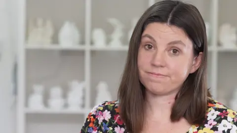 Jamie Niblock/BBC Sam Murray is looking at the camera with a half smile. she has shoulder-length dark brown hair and is wearing a flowered top. The picture is of her head and shoulders. 
