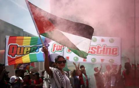 Getty Woman waving the Palestinian flag with a large banner saying Queers for Palestine in the background.