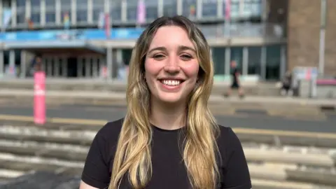 Young blonde woman looks into the camera and smiles. She is wearing a black t-shirt. In the background, you can see the main entrance to Swansea University's Singleton Campus.