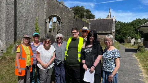 A group of people stand in front of a burned out church. They range in age from late teens to mid-70s. They are all dressed for sunny weather.