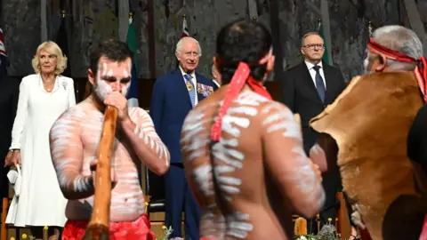 EPA Tiga pria mengenakan pakaian tradisional Aborigin, mengecat tubuh mereka dan bermain didgeridoo di bawah pengawasan Ratu Camilla, Raja Charles dan Perdana Menteri Australia Anthony Albanese