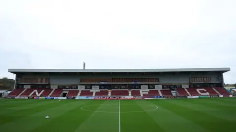 PA A wide shot if the Northampton Town football pitch and stadium. The stadium is currently empty with claret and white seats. 