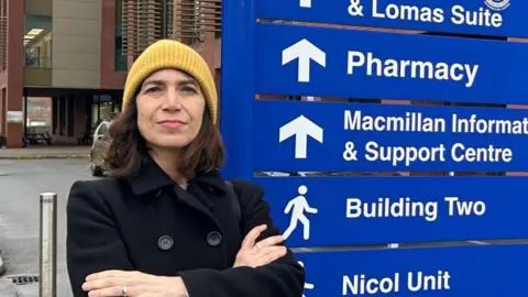 A woman wearing a black coat and yellow beanie hat stands in front of the Stratford Hospital sign. The sign includes directions and names of the various hospital departments and units.