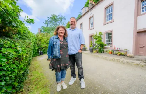 IWC Media Linda and Euan stand side by side on a gravel driveway in front of a light-coloured house with large windows and a doorway.
Lush green hedges line the left side, while potted plants and small garden features decorate the area near the building.
The scene is brightly lit under a partly cloudy sky, giving a vibrant and airy outdoor setting.