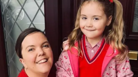 Family Fund Hanna is kneeling next to Darcy a the inside of a front door with a decorative lead frame window. Darcy has long blonde pig tail hair and is smiling for the cameral with a pink winter jacket and red jumper. Mum has long tied back brown hair and is smiling